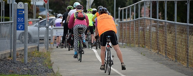 Group of people ride away from the camera on a concrete path towards a road, with the last rider wearing a high vis vest saying Bicycle Tasmania.