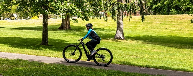 Woman rides up a path on an ebike surrounded by brilliant green grass and trees.