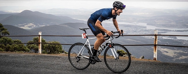 Man wearing blue lycra kit rides up slope on white road bike with a misty view of Hobart in the background.