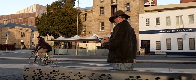 A man wearing a dark hat and coat looks down at a clipboard while a person rides past. Golden morning sun lights up the sandstone buildings in the background.