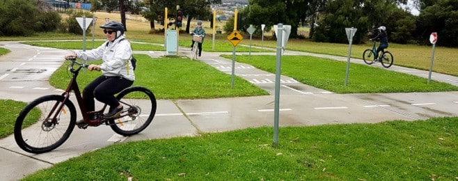 Three people ride on wet paths set up to look like a road network, with green grass inbetween the paths.