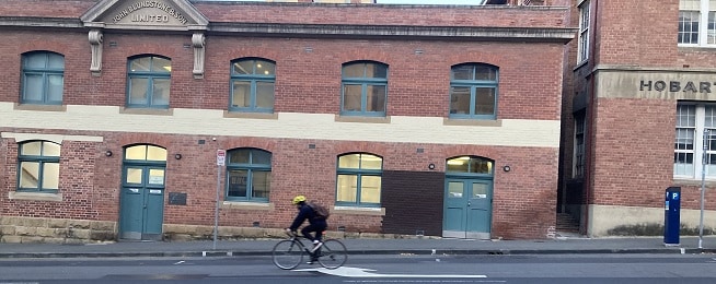 A man wearing a yellow helmet rides downhill in front of an old red brick building with a Blundstone sign at the top and Hobart lettering to the right, with green doors and windows.