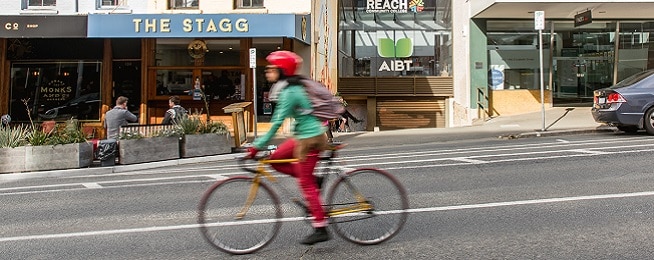 Woman wearing red pants and red helmet coasts down hill on Elizabeth Street on a classic road bike in front of the Stagg coffee shop.