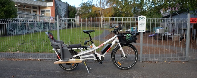 A white long-tail Yuba cargo bike with child seat and front basket stands alongside a fence with a 10 min parking limit sign and a please shut the gate sign, with a grassy lawn, building and trees on the other side of the fence.