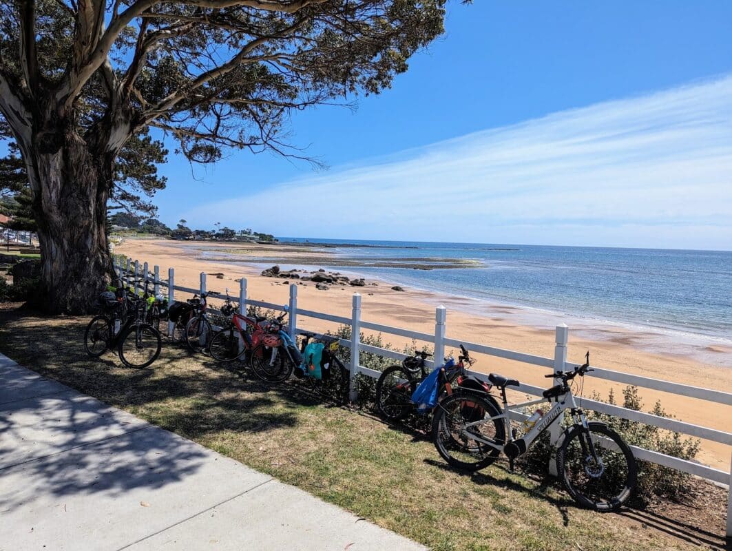 Bikes lined up along a white wooden fence next to concrete path with blue sky and beach beyond the fence.