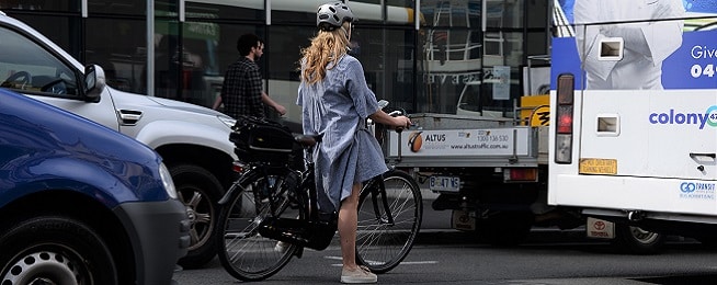 A woman with long blond hair wearing a loose chambray dress stands astride her stepthrough ebike on a road inbetween a bus and car.