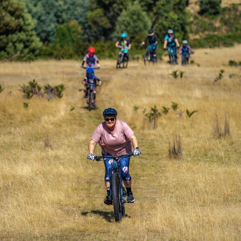 An older woman rides across a grassy field with other riders following.