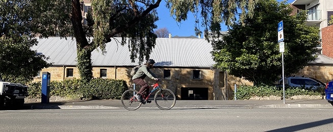 Man rides along Collins Street in Hobart with a sandstone building and trees in the background.
