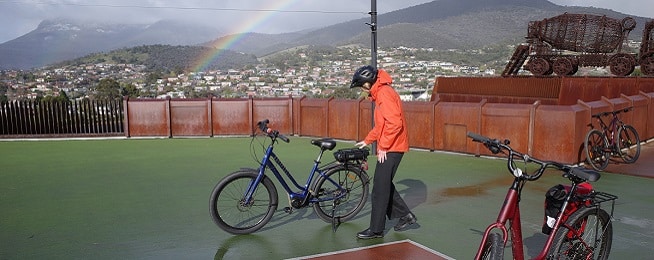 A person in an orange jackets and black pants and bike helmet inspects a blue ebike in the forecourt at MONA with a grey sky and rainbow in background.