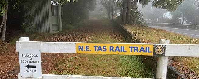 A yellow sign with blue lettering saying N.E. Tas Rail Trail on a white wooden fence with a foggy bush backdrop.