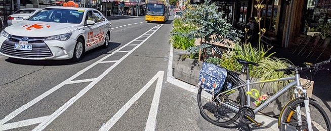 Narrow painted bike lane on a street with a bus and taxi on one side and plants and parked bike on the other.