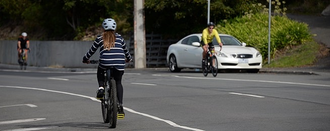 A teenager rides along Sandy Bay Road with two road cyclists in the distance on the opposite side of the road.