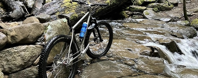 Silver single speed mountain bike sits on rock shelf with water flowing over it and bush in the background.