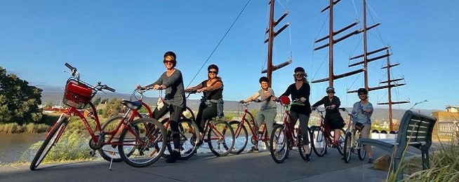 A group of women on bikes sit on asphalt path with ship masts and blue sky in the background.