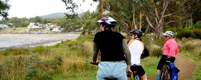Three women sit on bikes on a dirt trail looking out over the water.