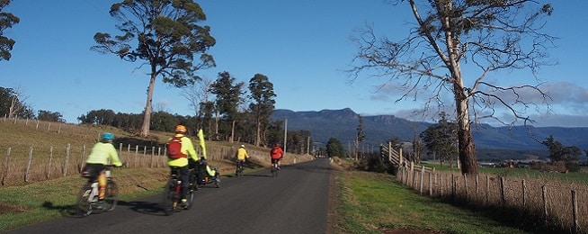 A group of riders wearing brightly coloured jackets on a road towards a mountain range flanked by farmland and fences.