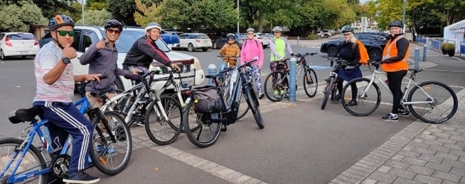 A group of people with bikes stand in a line next to a car park, with some waving at photographer.