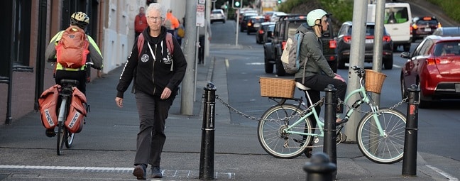 A woman holding a pale blue bike and wearing a matching helmet waits to cross Davey Street in Hobart , woman wearing a black hoodie walks towards the camera and a woman rides away from the camera with two loaded worn-out panniers and backpack.