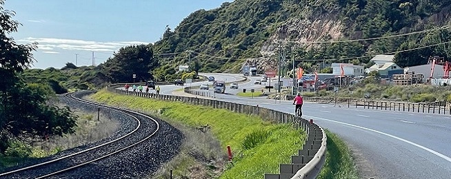 A road curves around a coastline with rail lines on the left and high cliffs to the right, a group of people are riding on the far left of the road.