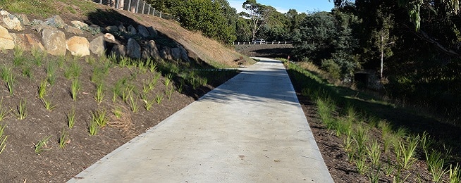 A newly laid concrete path runs along the side of a hill with new plantings on the left of it and a stream at the bottom of the hill to the right, a bridge with a car on it can been seen in the background.