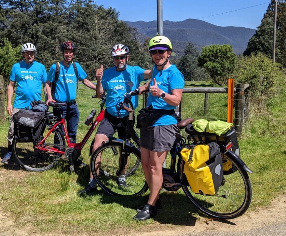 Four people stands with bikes on a the side of a rural ride all wearing blue tshirts with "What Hills?" and a symbol of a bicycle on the front.